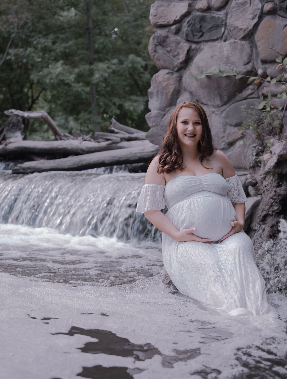 A serene maternity portrait taken near a rocky waterfall in white lace dress.