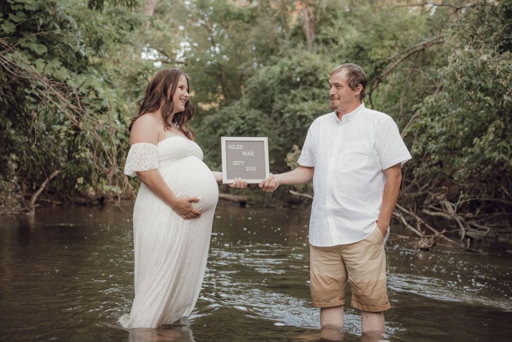 Couple standing in stream holding letterboard for pregnancy announcement.