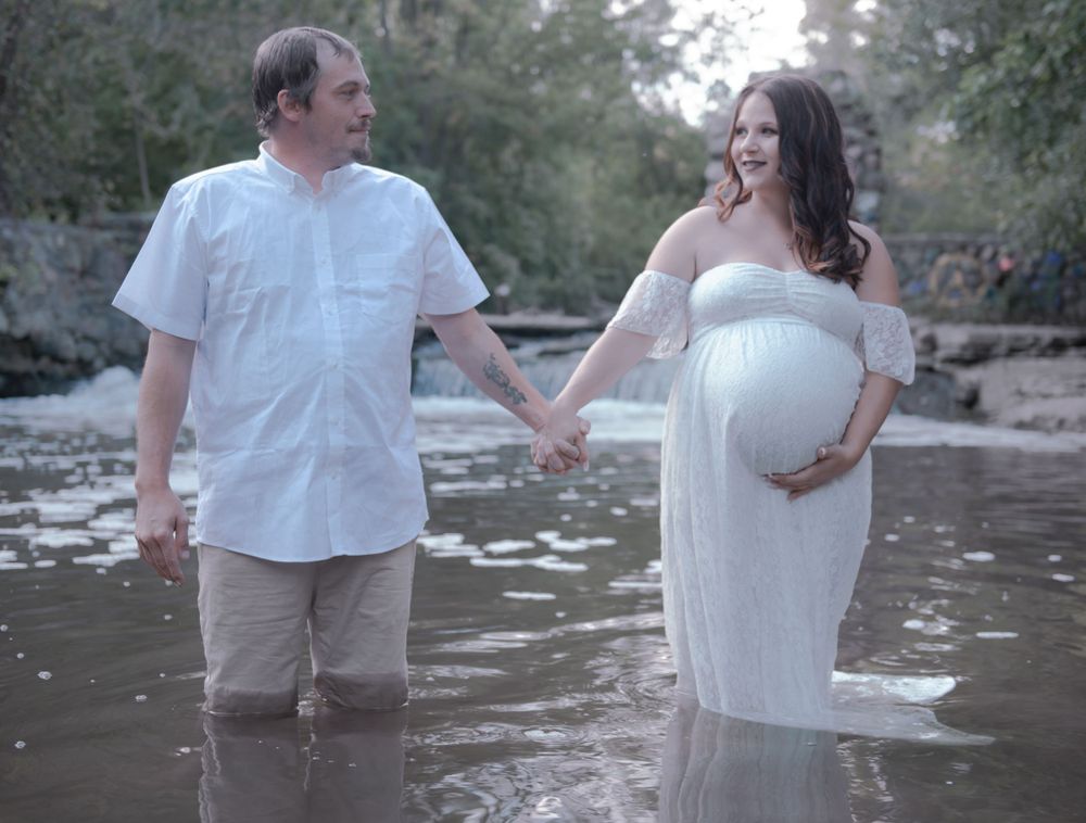 A maternity photoshoot with a couple standing in shallow water wearing white.