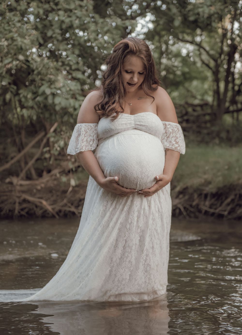 Ethereal maternity photo in flowing white dress standing in shallow stream water.