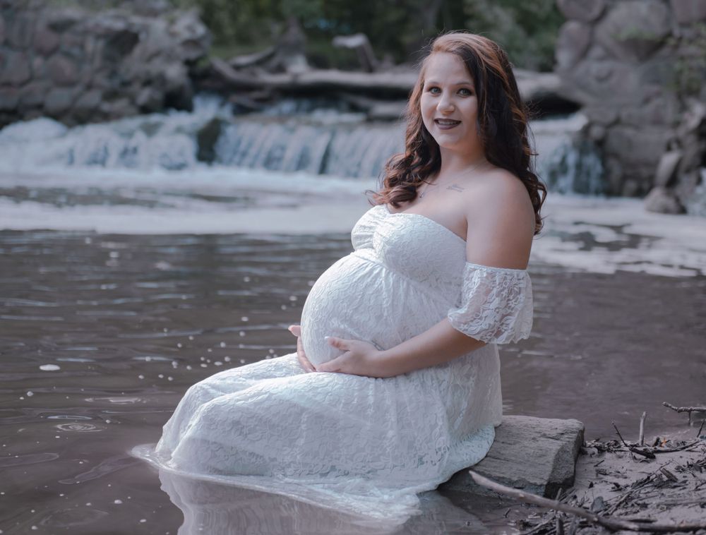 A maternity portrait of an expectant mother in a white dress by a waterfall.