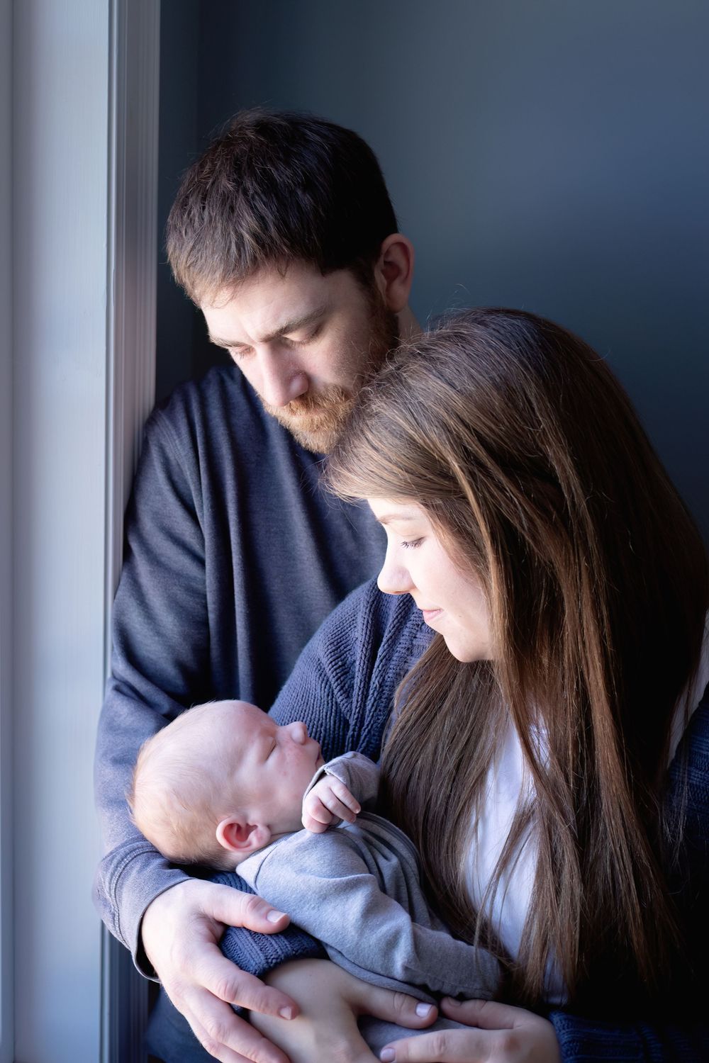 Family shares tender moment while holding newborn baby by window with natural light.