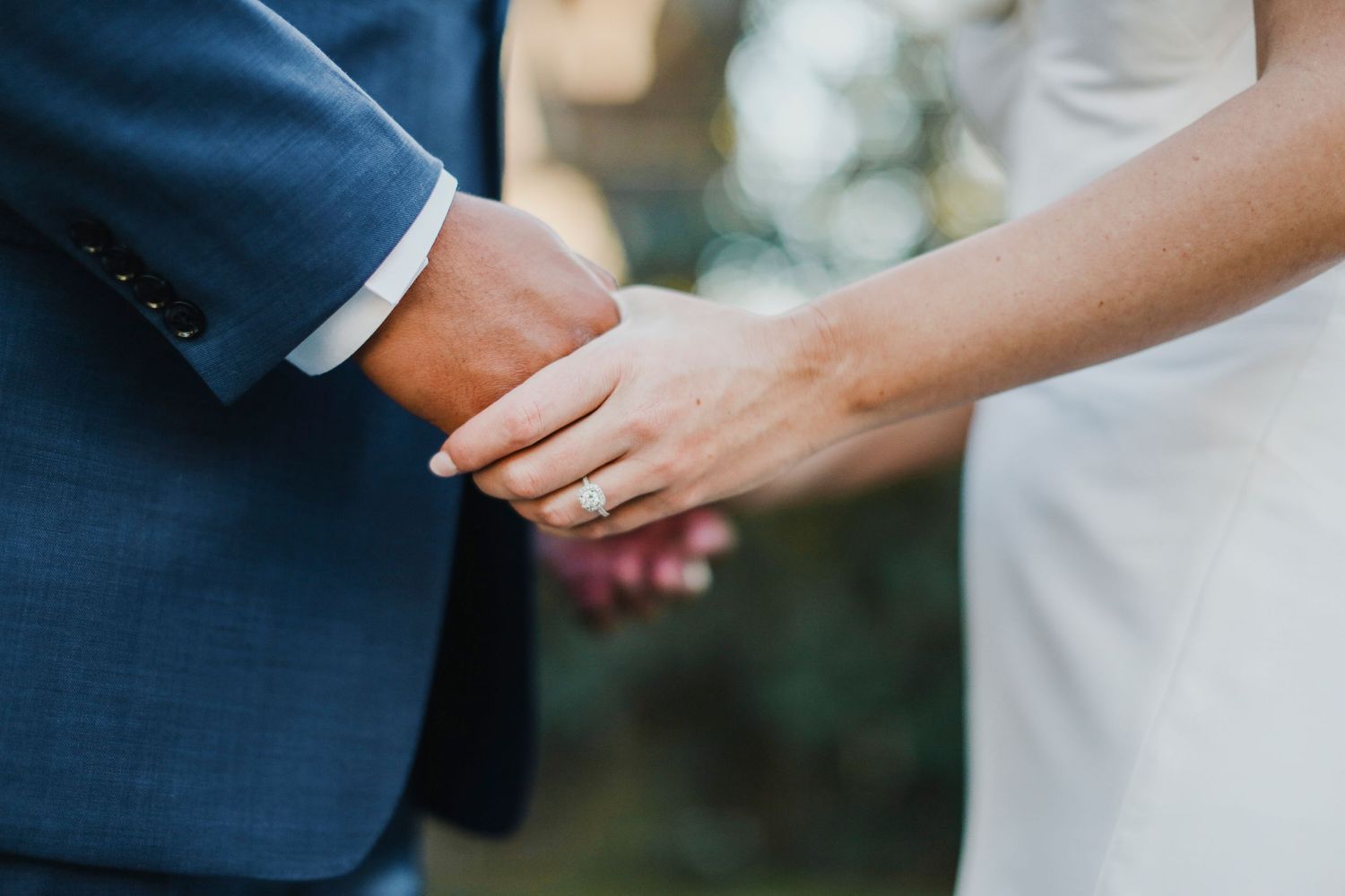 A close-up of hands in tender embrace during a wedding ceremony, with navy suit sleeve and white dress details visible.