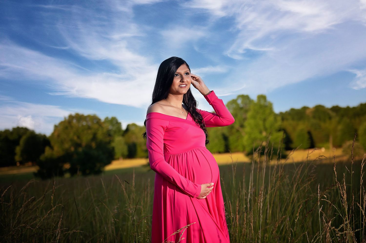 A maternity portrait in a bright pink dress against a golden sunset in an open field.