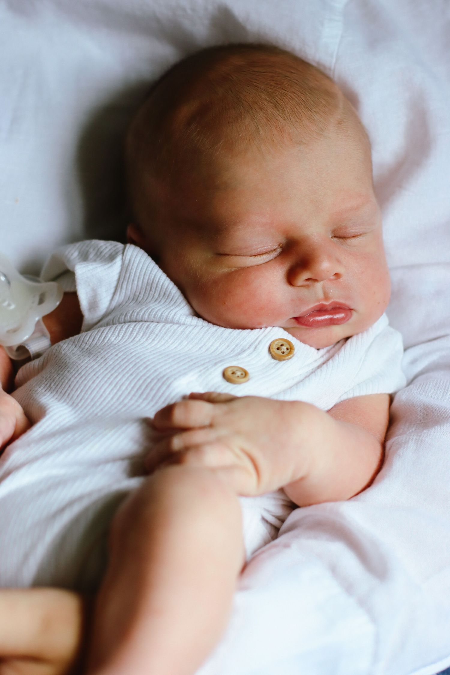 Sleeping newborn wearing white buttoned onesie on soft bedding.