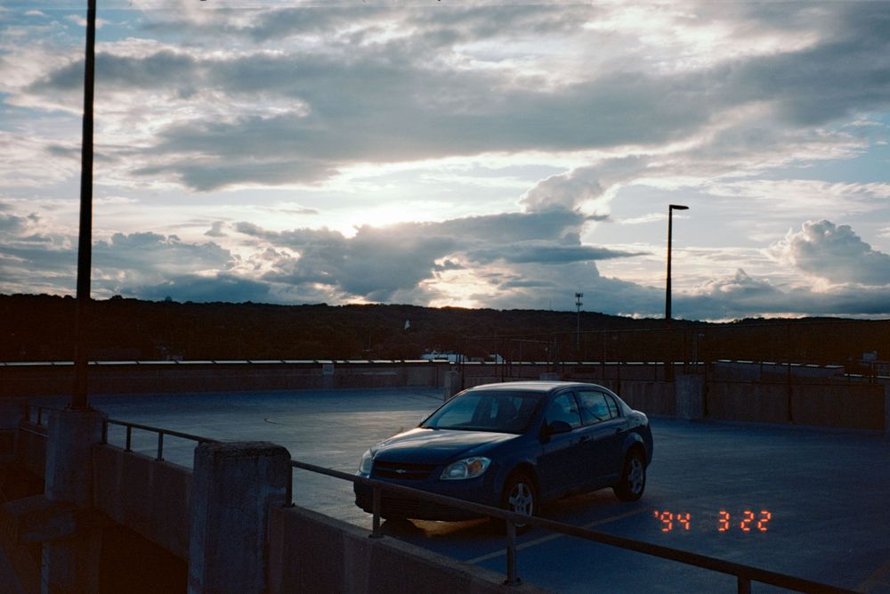 Blue sports car parked in lot during sunset with dramatic cloudy sky.