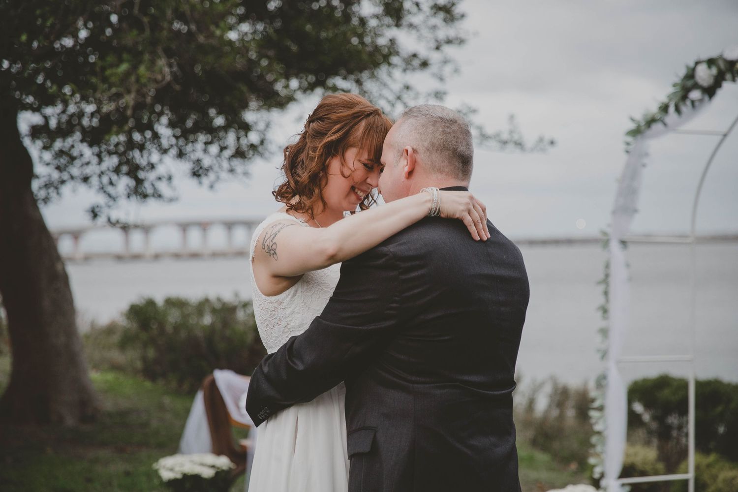 Bride and groom share a tender embrace during outdoor waterfront wedding ceremony under floral arch.