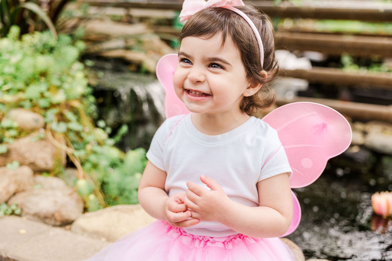 Joyful toddler in fairy costume with pink wings plays in garden setting.