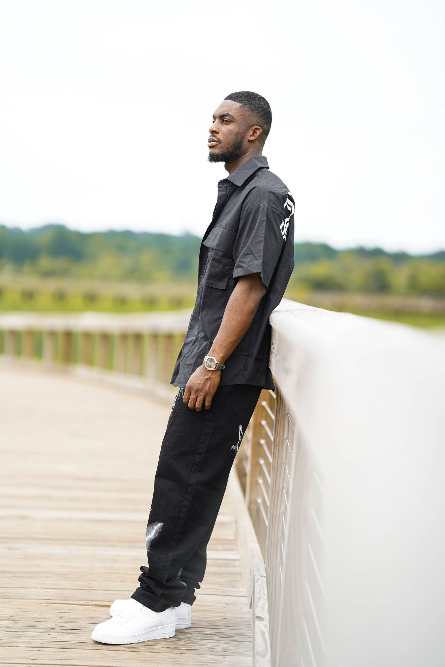 Fashion model in all black attire poses casually on wooden boardwalk overlooking natural landscape.