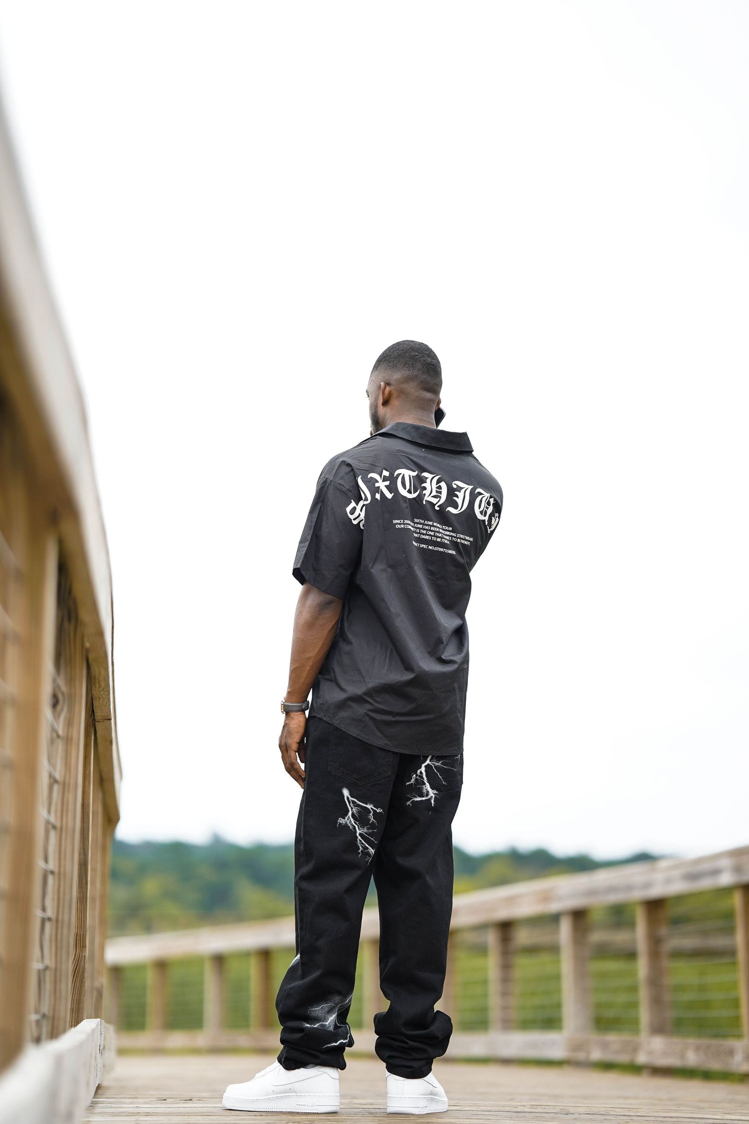 Model wearing black clothing poses from behind on wooden walkway with scenic background.