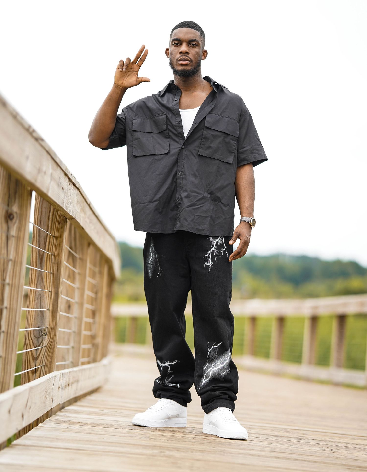 Fashion model in black streetwear poses confidently on wooden boardwalk during outdoor photoshoot.
