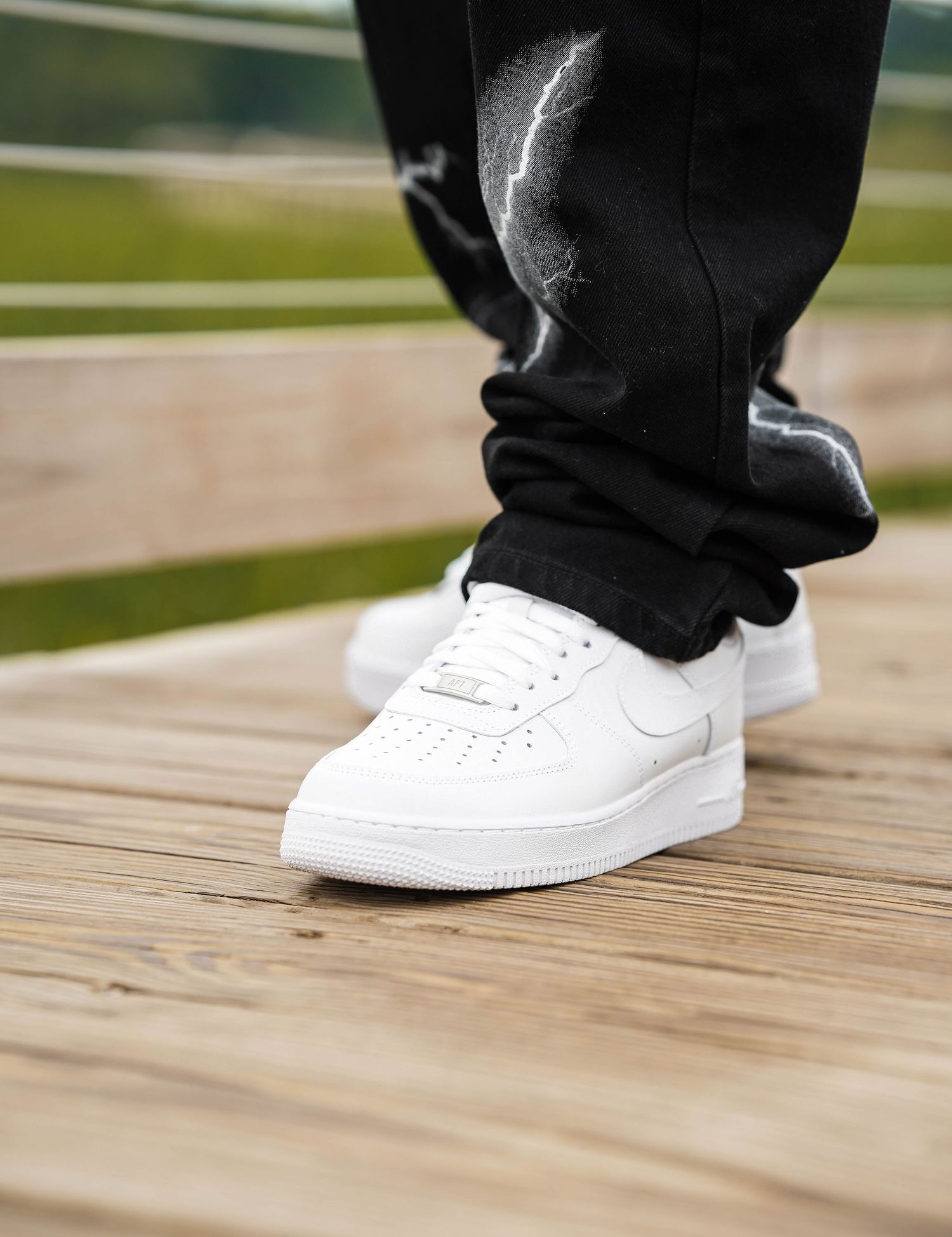 Detail shot of white sneakers and black pants on wooden boardwalk during fashion photoshoot.