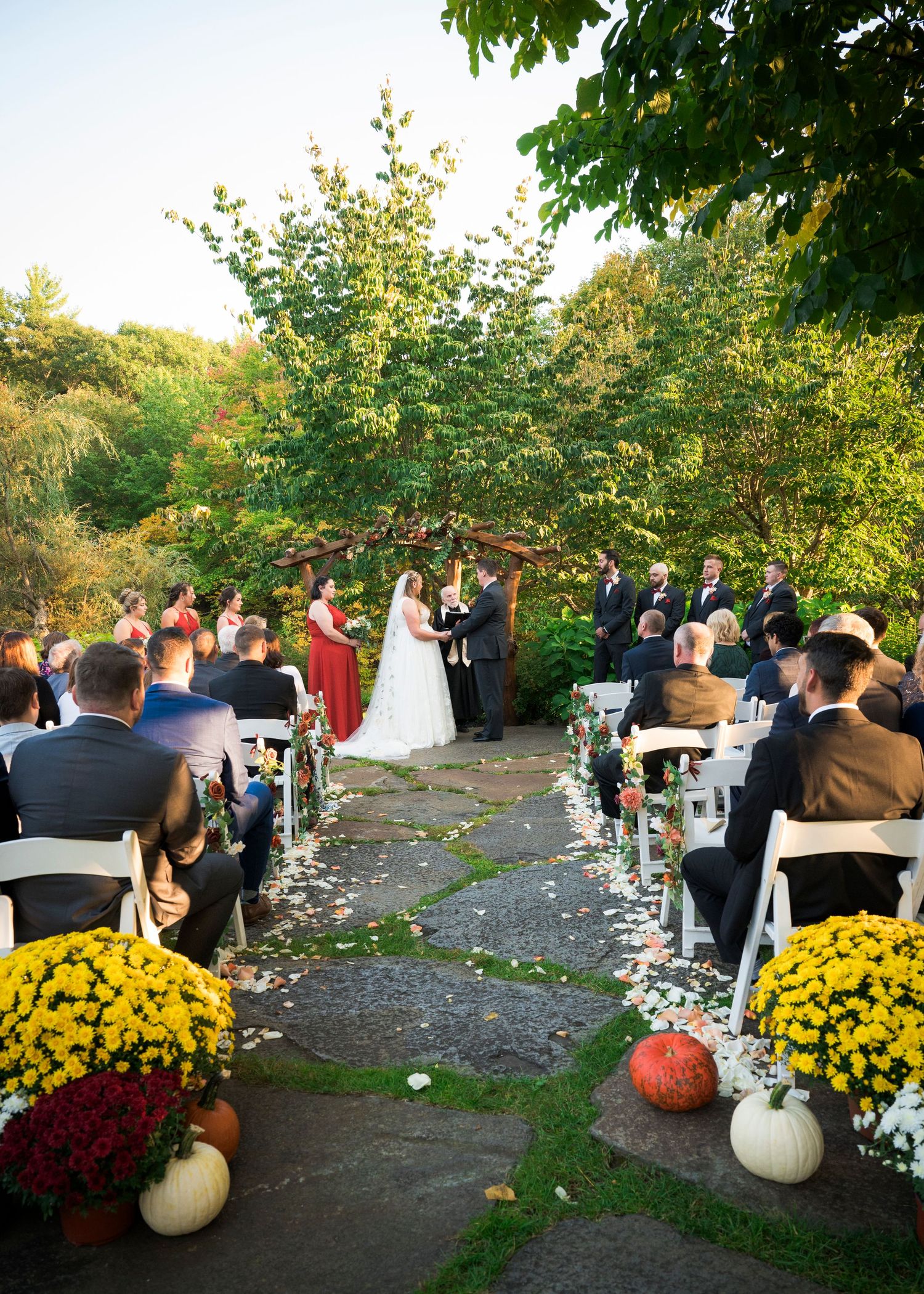 An outdoor wedding ceremony with rows of white chairs and autumn decor featuring pumpkins and yellow mums.