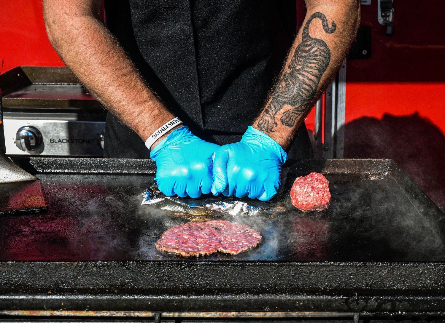 Chef wearing black with blue gloves grilling meat patties on a flat top griddle.