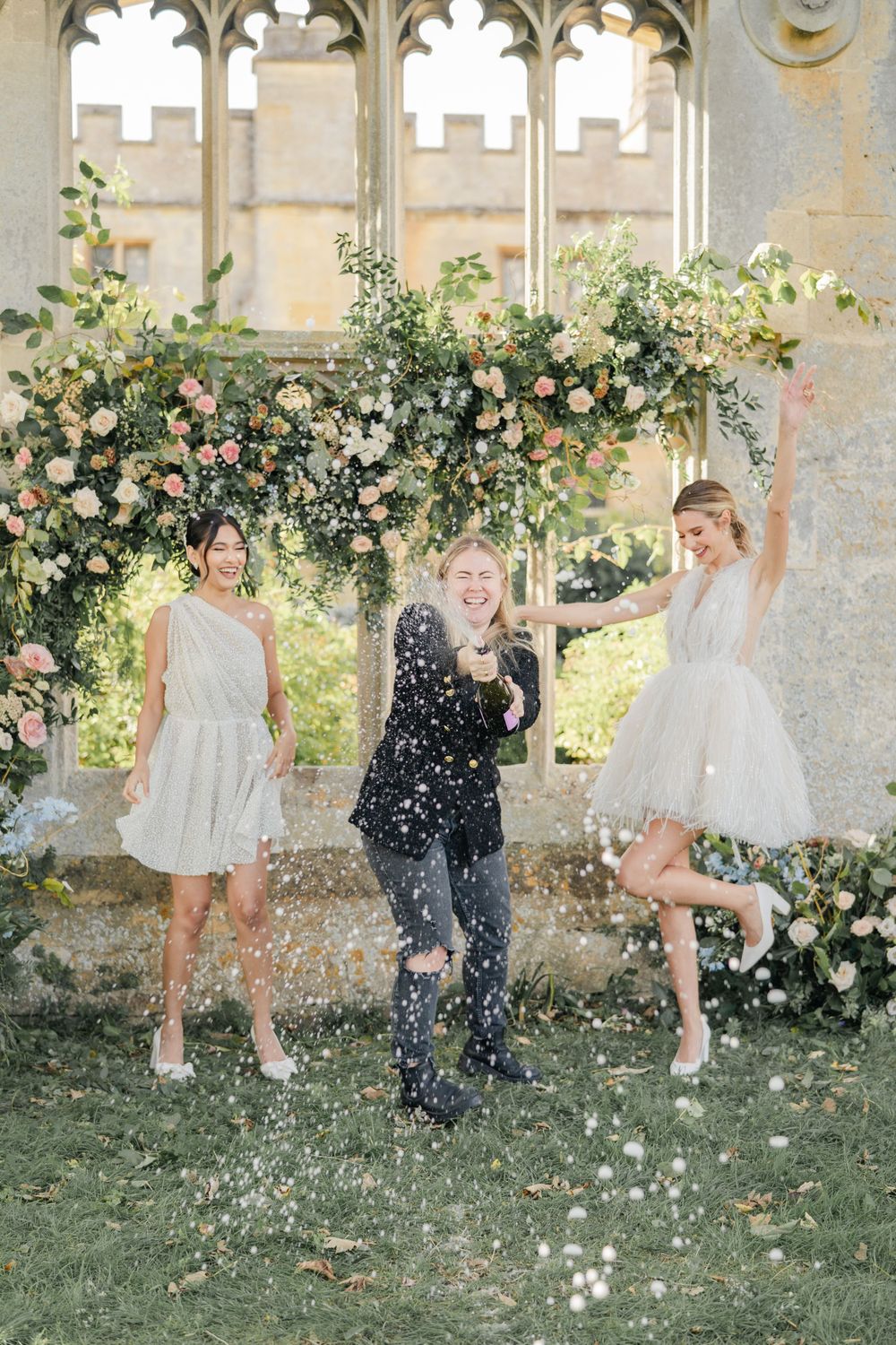 Joyful wedding party throws flower petals in a garden setting with gothic architecture.