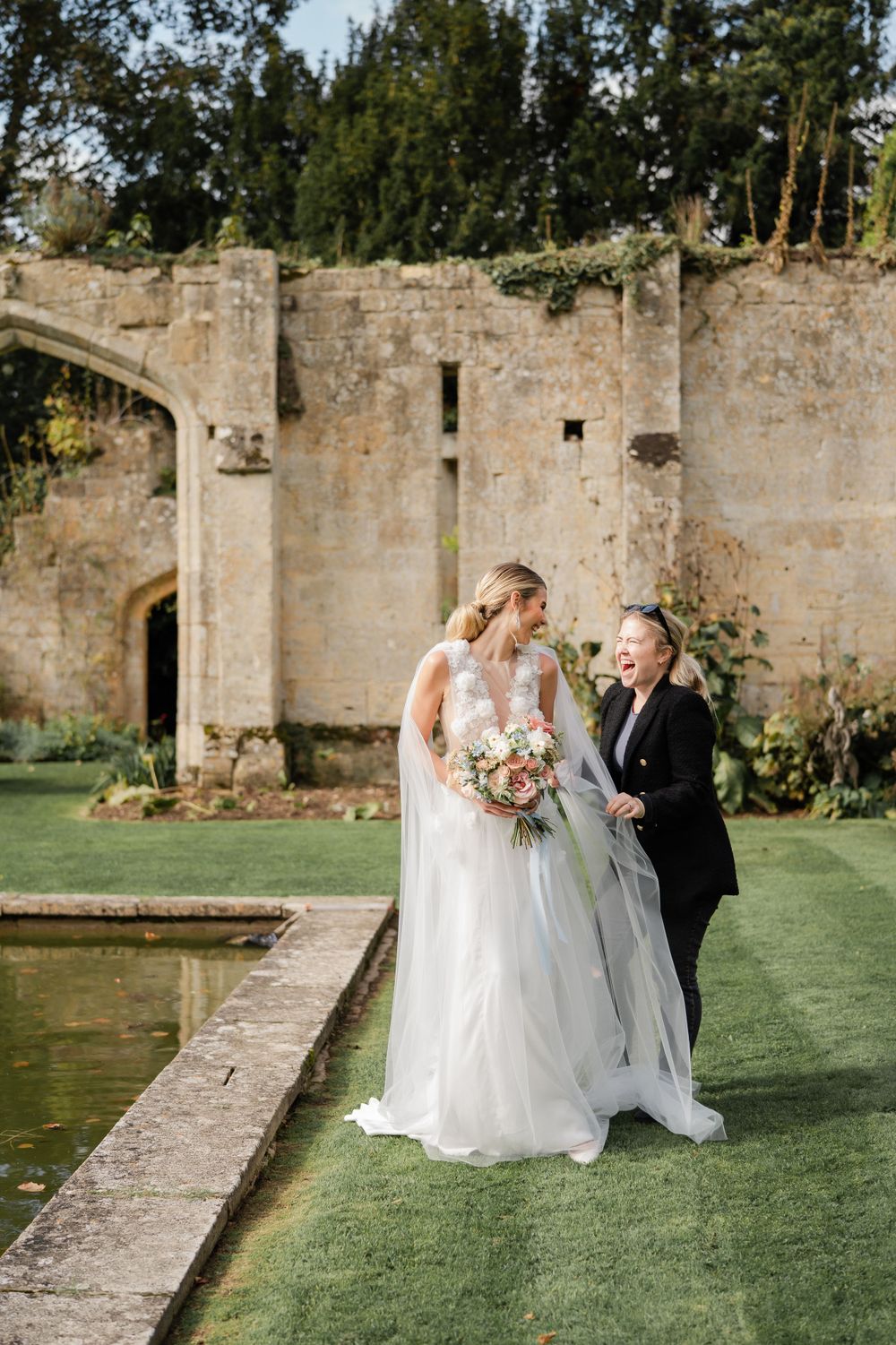 Wedding party poses for photos near historic stone walls and a reflective pond.