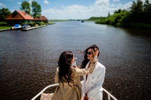 Two figures share a moment on a boat with a scenic river and waterfront houses in the background.