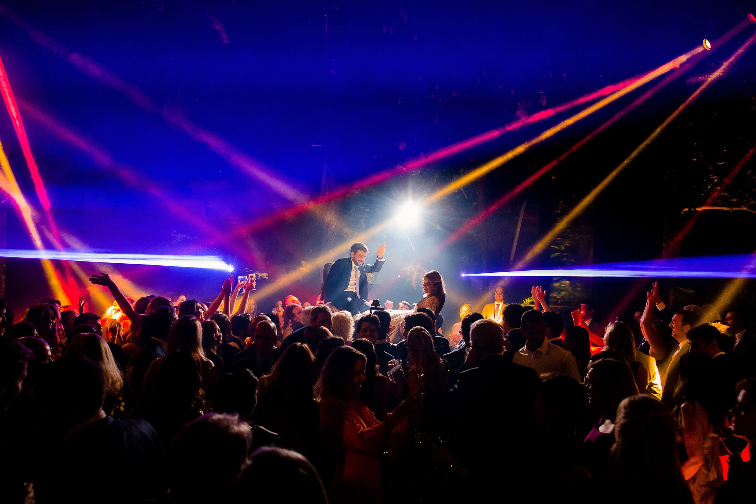Colorful laser lights illuminate a crowded dance floor during a nighttime celebration.