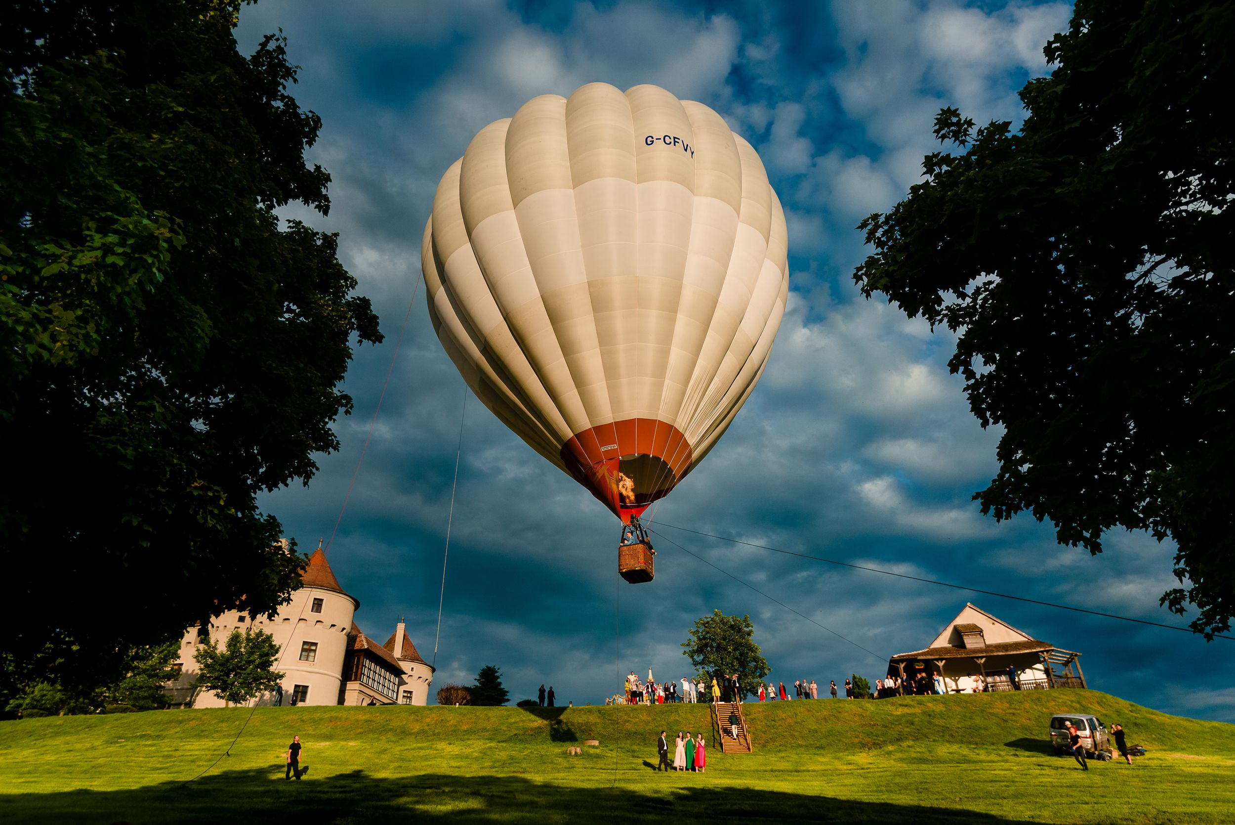 Hot air balloon floats over a green hillside with historic buildings in the background.