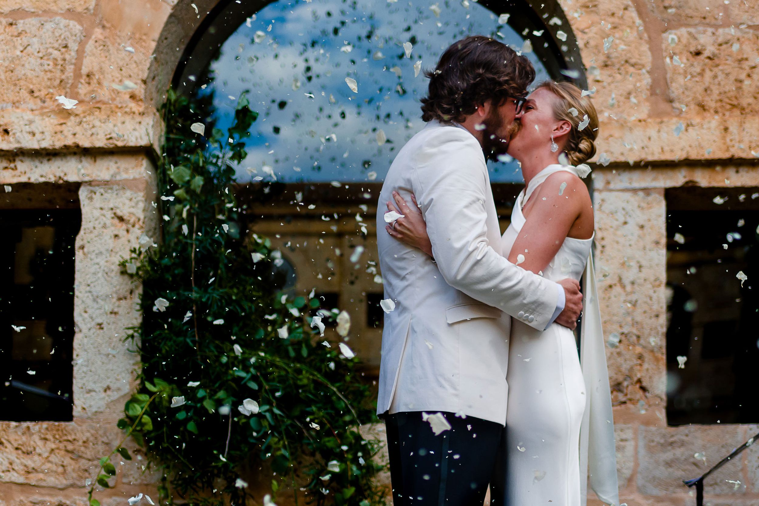Romantic couple embraces under a stone archway as confetti falls around them during a wedding celebration.