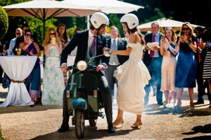 Couple departing wedding on vintage scooter while guests wave goodbye under umbrellas.