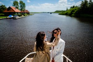 Two people share an intimate moment aboard a boat on a serene river with rustic houses visible along the shore.