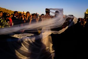 A flowing white veil catches the wind during an outdoor celebration surrounded by onlookers at sunset.