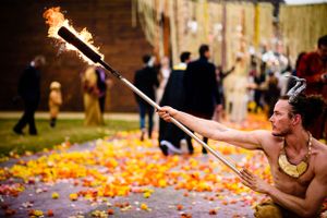 A fire performer swings a flaming staff in front of a dramatic backdrop covered in orange flower petals.