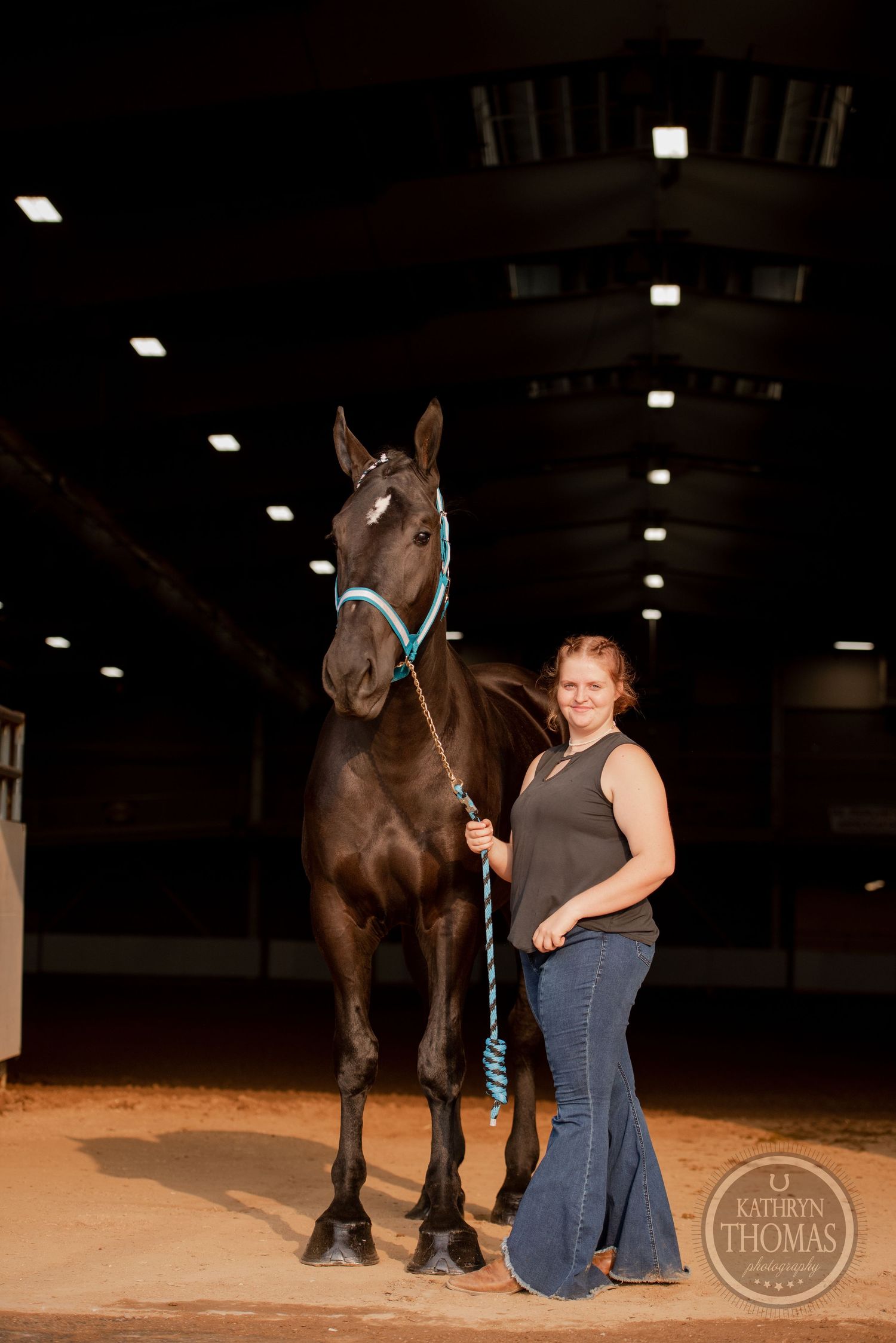 A dark horse with turquoise halter stands alongside its handler in an indoor equestrian facility.