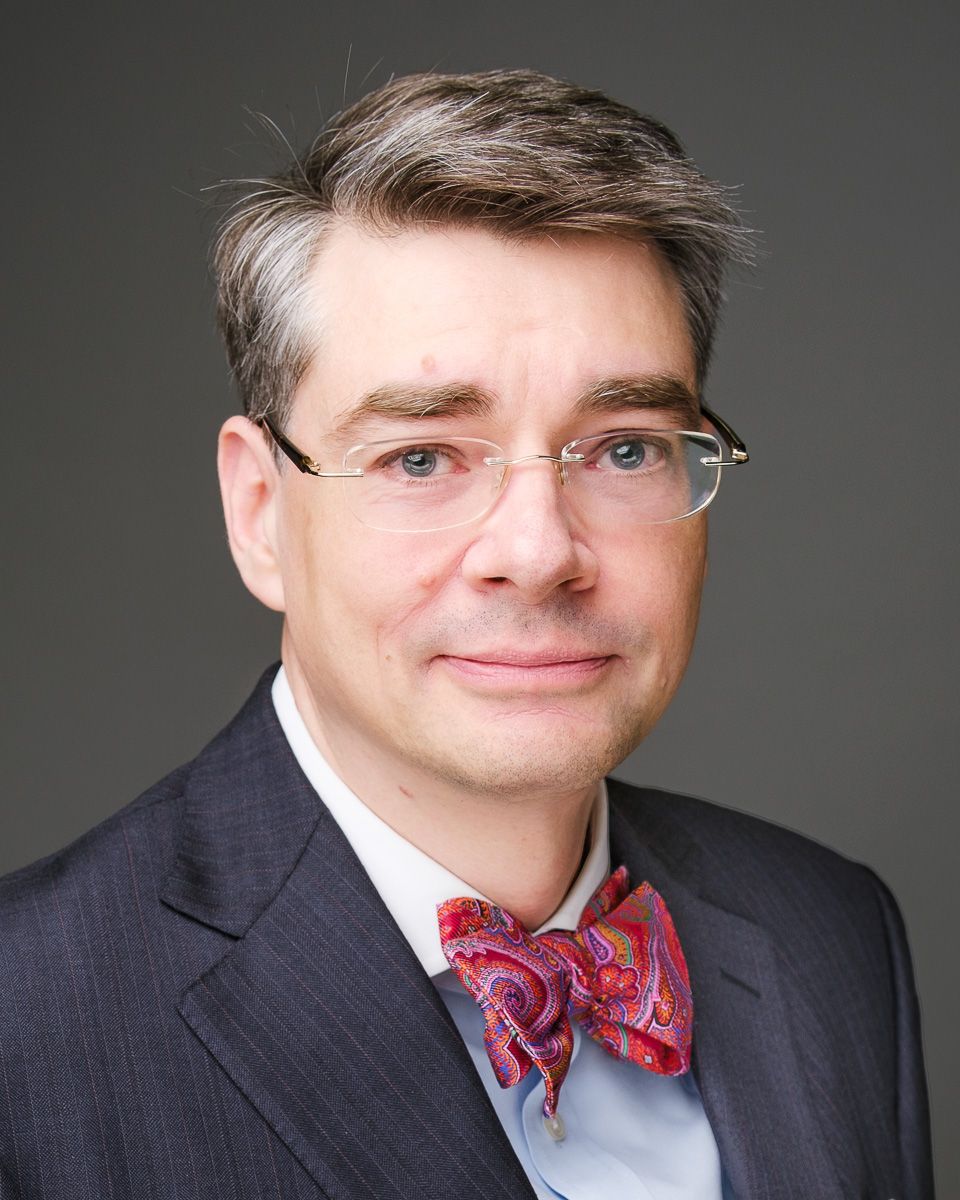 Professional headshot of someone wearing glasses, a navy suit jacket, light blue dress shirt and patterned red bow tie.