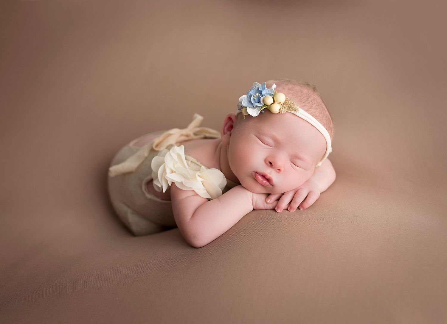 Sleeping newborn wearing a delicate floral headband and cream-colored wrap on brown backdrop.