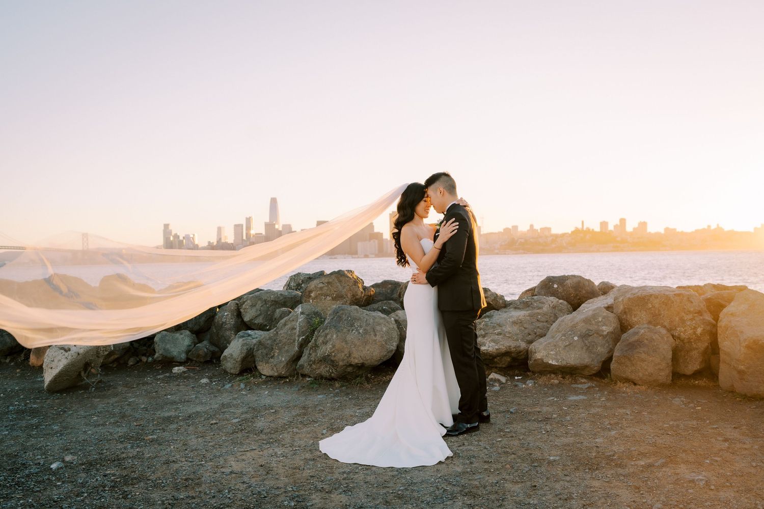 A wedding couple shares a romantic moment on rocky terrain as the bride's veil flows in the wind at sunset.