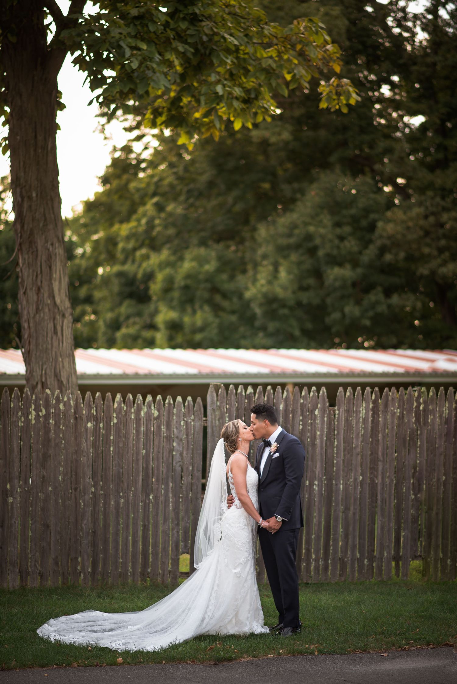 Bride and groom kissing in front of a wooden fence surrounded by lush green trees on their wedding day.