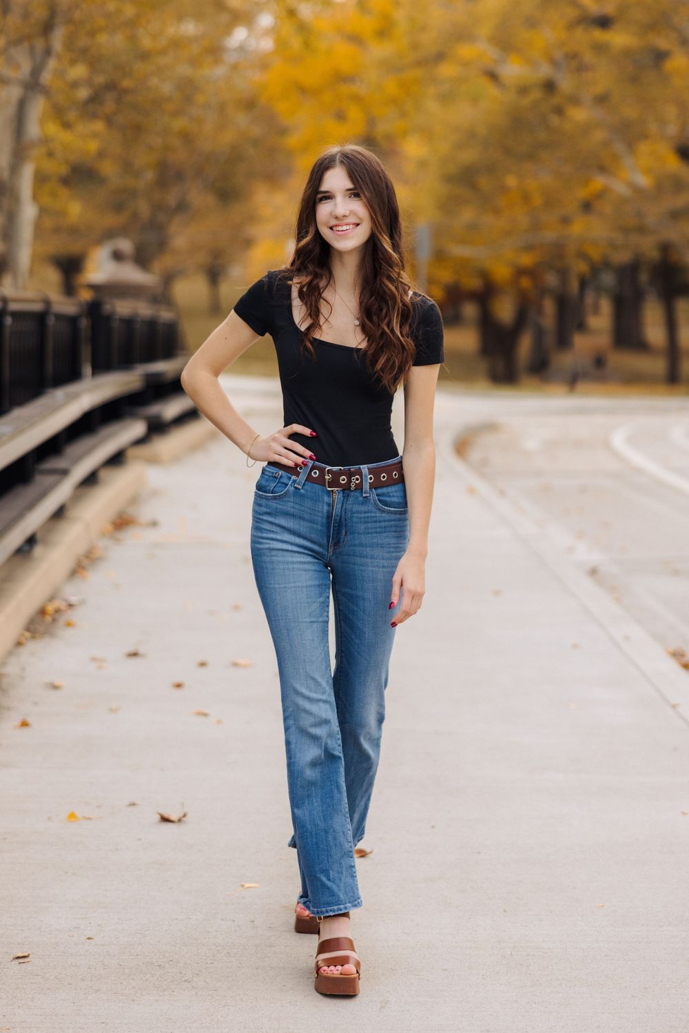 Portrait of person in black top and jeans walking on path with autumn foliage in background.
