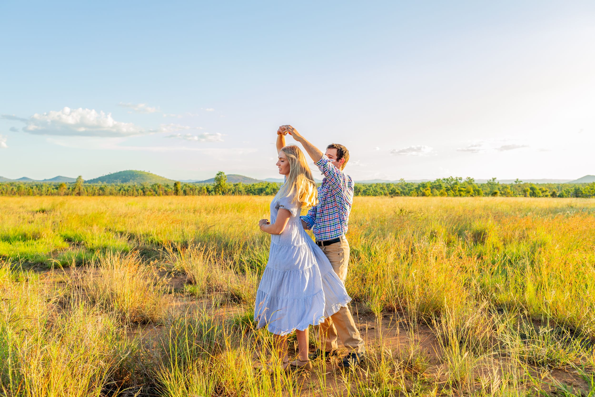 A couple dances joyfully in a golden meadow with mountains and blue sky in the background.