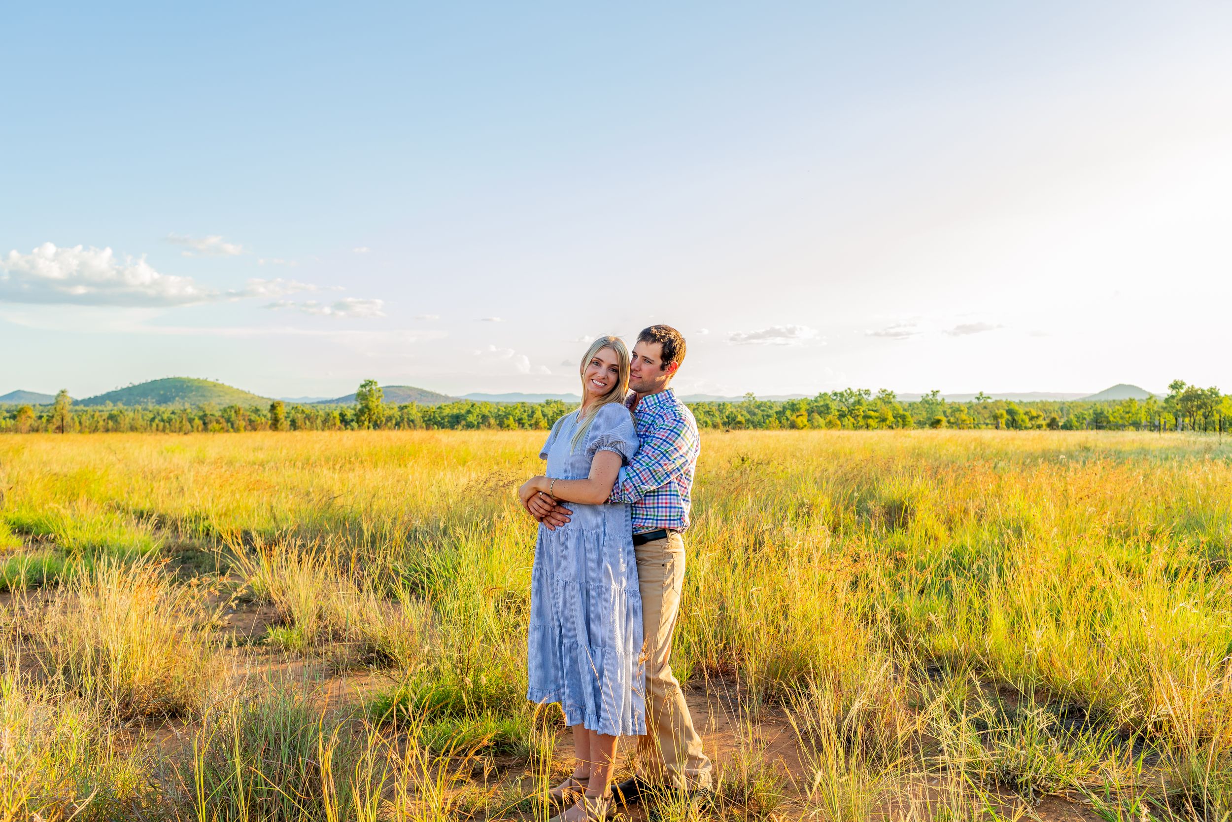 A romantic portrait in a sun-lit field with mountains and blue sky creating a dreamy backdrop.