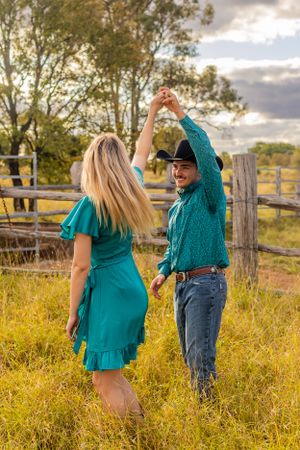 A couple in matching turquoise outfits dance together in a field with wooden fencing at golden hour.