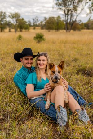 A couple and their red cattle dog pose together in a golden field during sunset.
