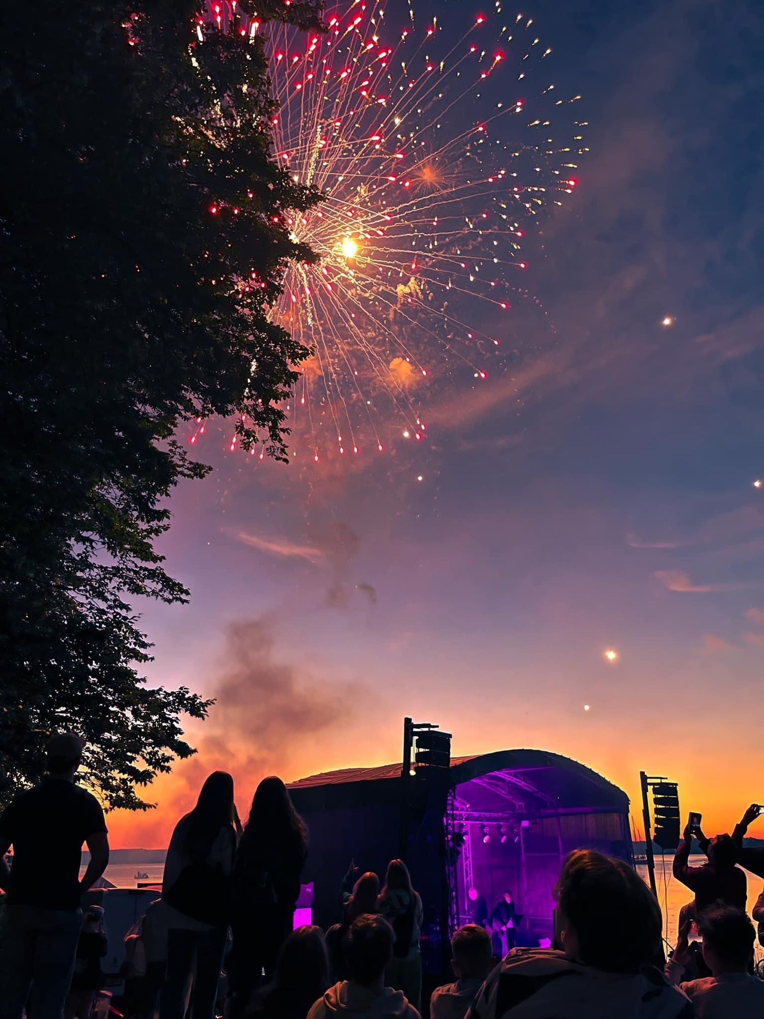 Colorful fireworks display lighting up the night sky above a festival crowd with purple stage lights.