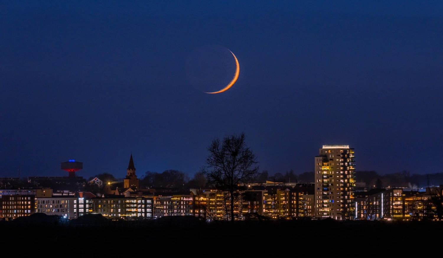 City skyline at dusk with a crescent moon visible in the dark blue sky.