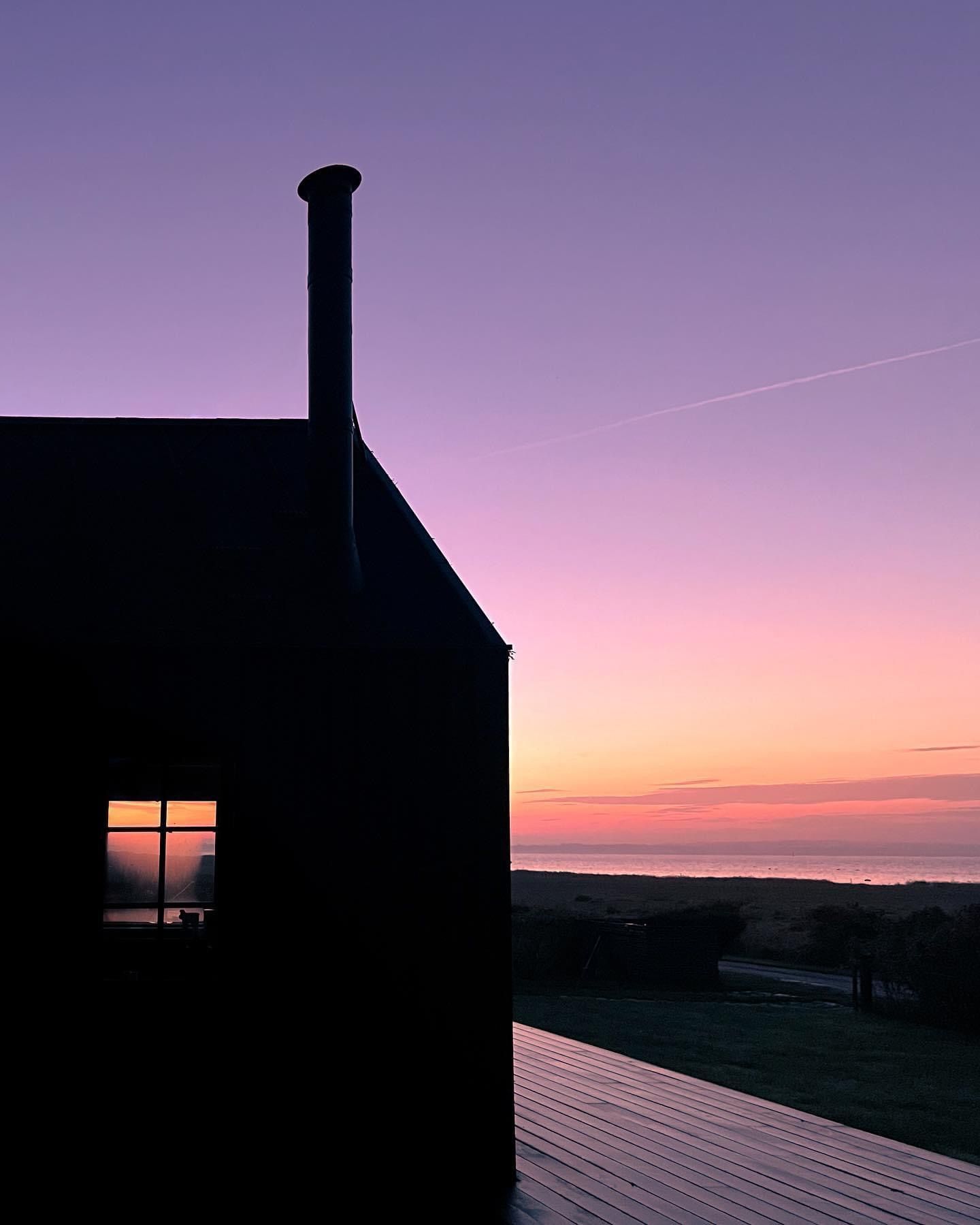 Silhouette of house with illuminated window against purple and orange sunset sky.