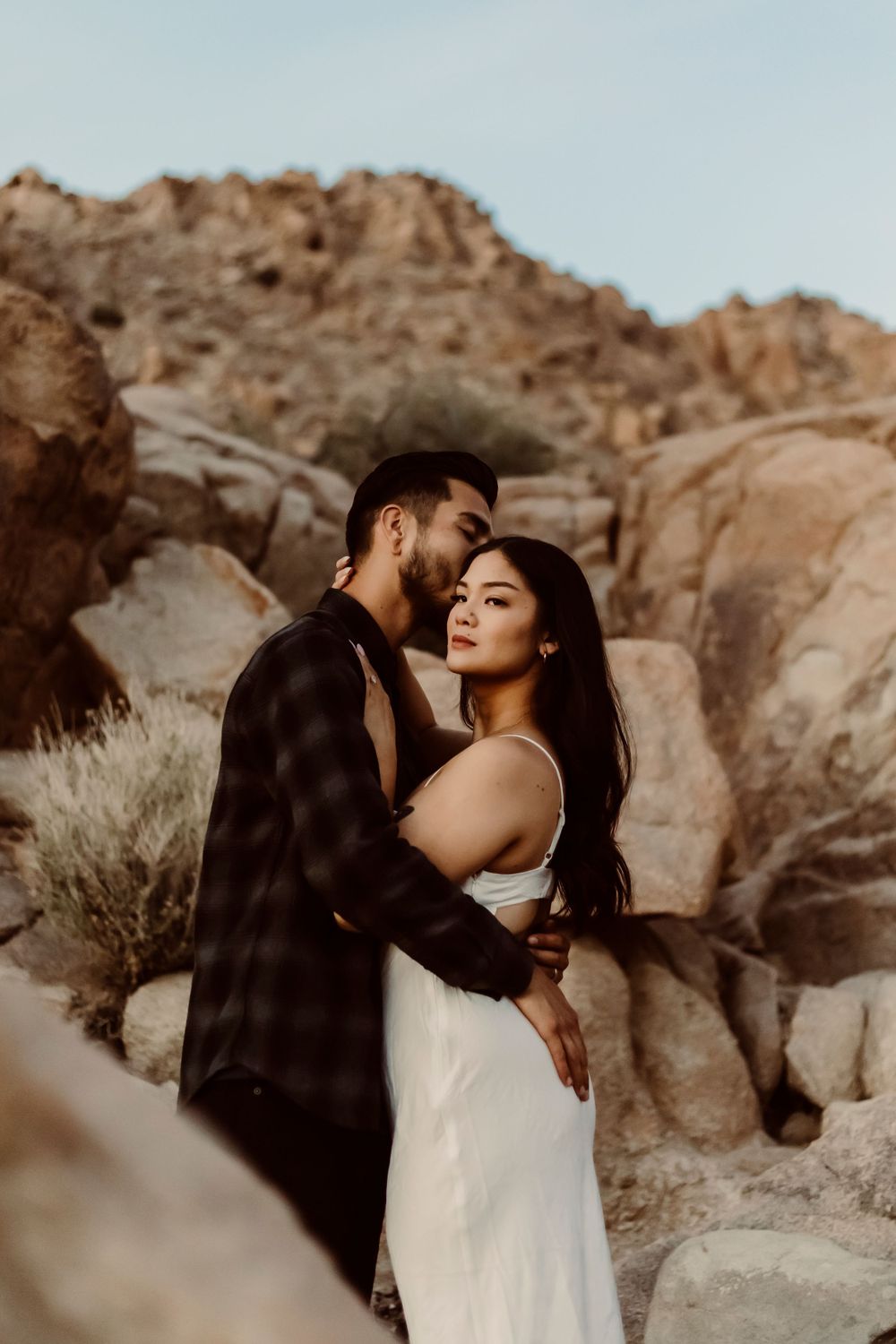 A romantic couple embraces among Joshua Tree desert rock formations during a golden hour photoshoot in nature