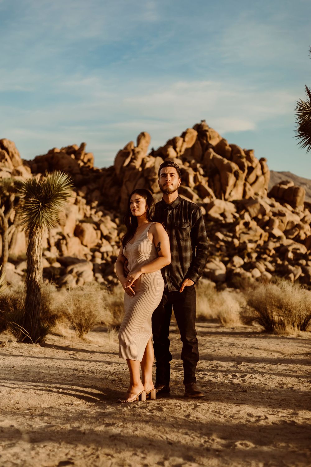A couple stands together in the desert landscape of Joshua Tree National Park with large rock formations in the background