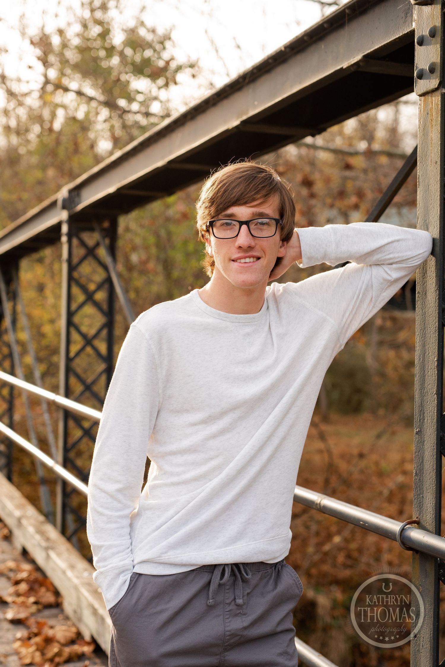 A person in a white sweater poses against metal bridge supports in fall scenery.