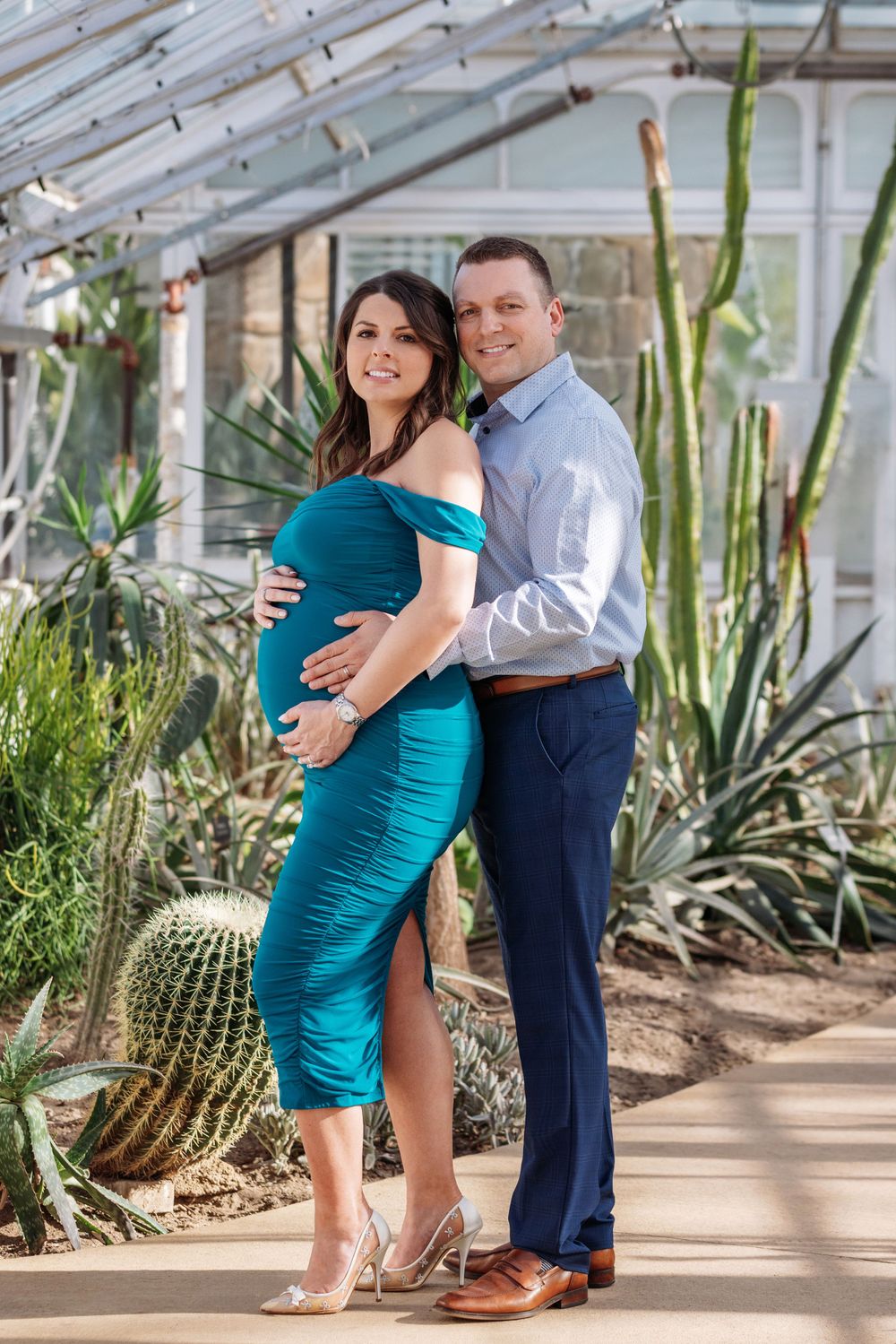 A couple poses in a greenhouse surrounded by cacti and desert plants as they embrace for a maternity photo.