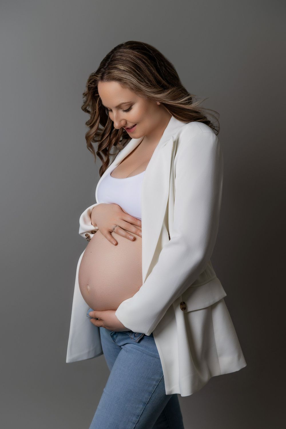 Expectant mother in white cardigan and jeans tenderly cradles her baby bump against a gray studio backdrop.