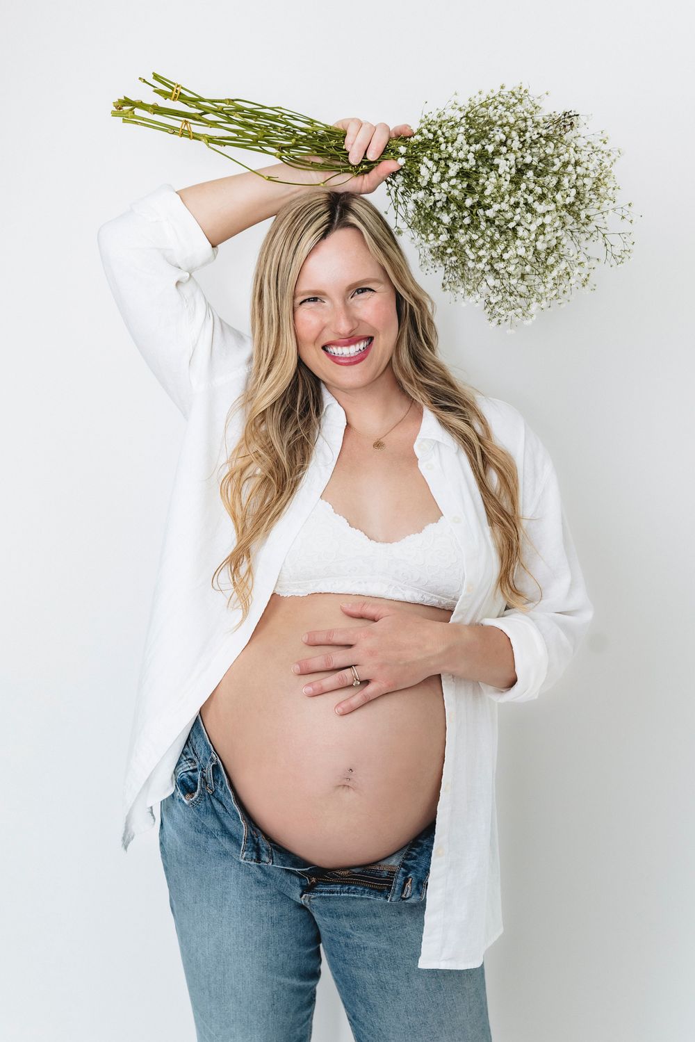 Expectant mother in white shirt and jeans holds baby's breath flowers against white background while touching her belly.