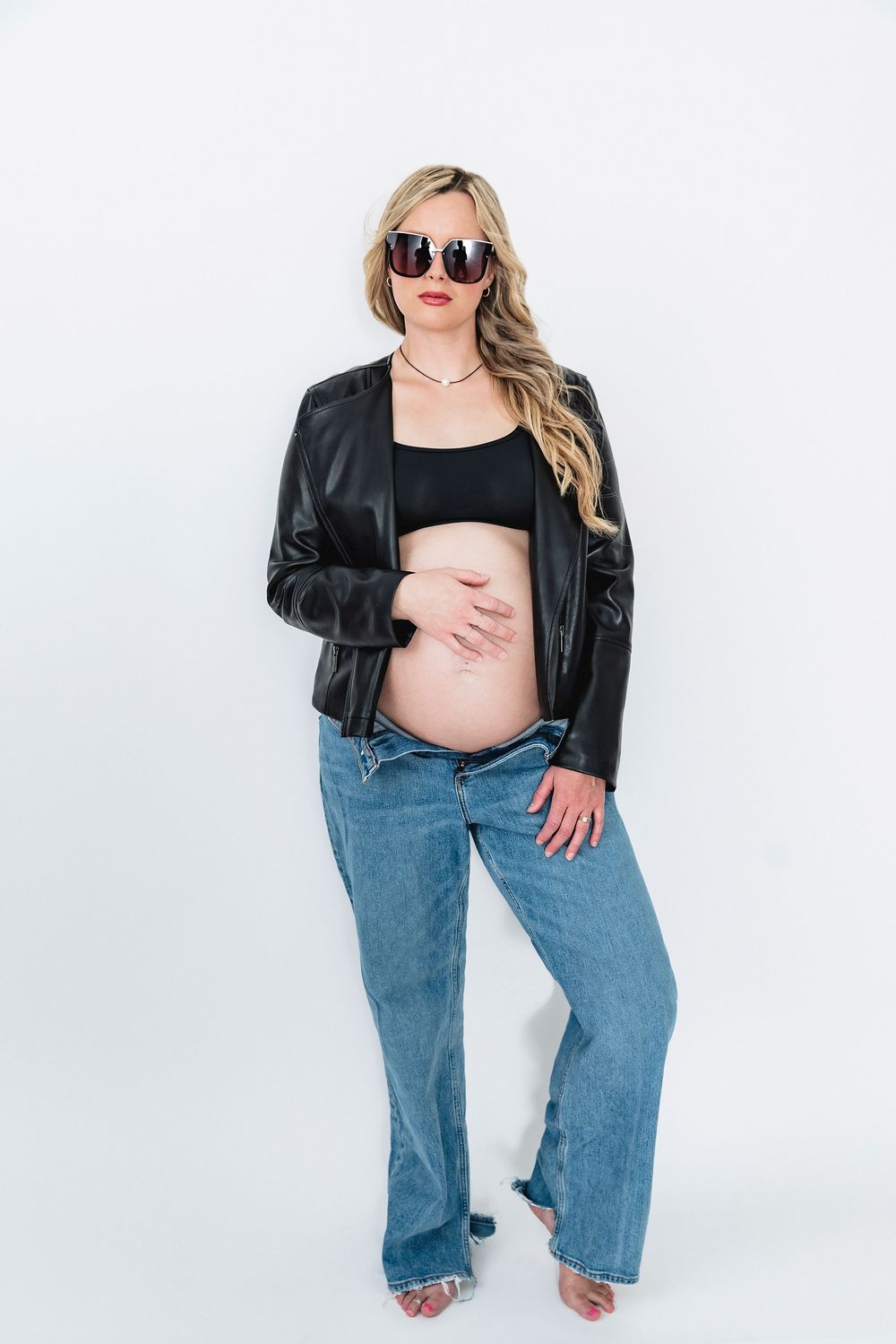Pregnant woman in leather jacket and jeans poses against white background wearing sunglasses and showing baby bump.
