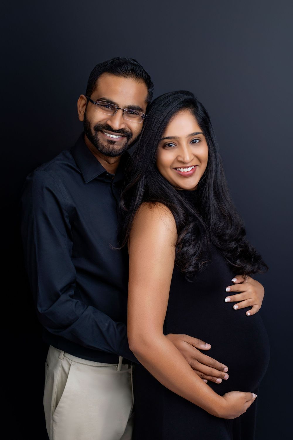 A couple poses together against a dark background in matching black outfits for an intimate portrait session.