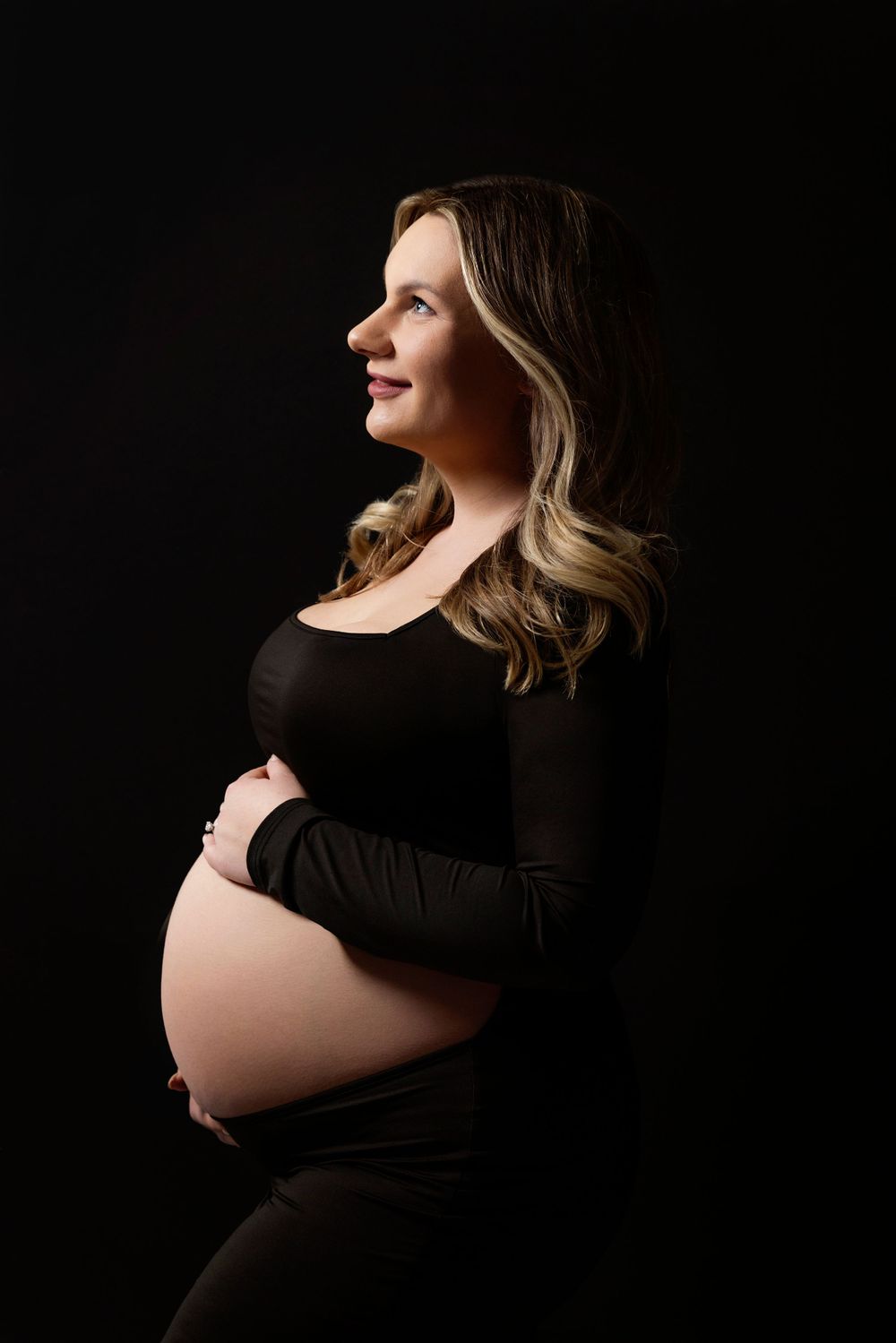 Classic maternity portrait of an expectant mother in black dress against dark background, photographed in profile.
